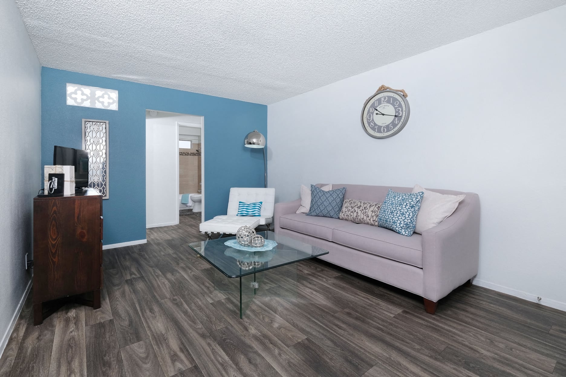 Modern living room in fifteen 50 apartments Las Vegas with grey plank tile floor, grey sofa, glass coffee table, and blue accent wall.